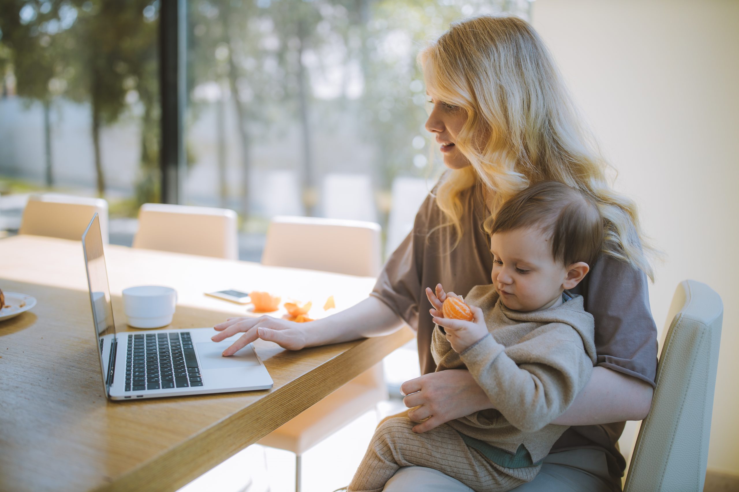 woman at table on laptop holding child eating an orange
